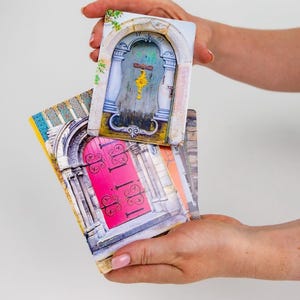 Puede incluir: Una pila de posavasos rectangulares con imágenes de puertas coloridas. Un posavasos muestra una puerta turquesa con un acento amarillo, mientras que otro muestra una puerta rosa vibrante. Los posavasos están sostenidos en las manos de una persona.