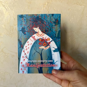 May include: A deck of "Manifestation" metaphoric associative cards. The cover art depicts a woman in a white embroidered dress with red floral details, surrounded by birds and a tree with red berries, against a blue backdrop.