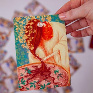 May include: A tarot card illustration featuring a woman with long red hair and a white robe. Her hair flows into a pool of dark liquid with green leaves. The card has a teal and gold background.