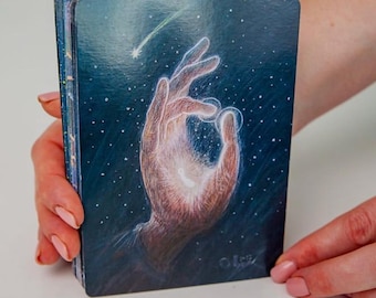 Peut inclure: Un jeu de cartes oracle avec une main tendue vers une lumière brillante dans un ciel étoilé. Les cartes ont un fond bleu foncé avec une étoile filante. Le jeu est tenu par une personne.