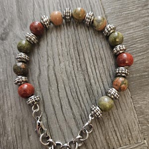 May include: A beaded bracelet featuring a mix of red, green, and tan-colored round beads. The beads are separated by silver-colored metal spacers. The bracelet has a silver-colored chain and clasp closure.
