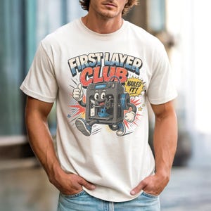 May include: Cream-colored t-shirt featuring a cartoon 3D printer with the text "FIRST LAYER CLUB" in blue and red. The printer character is giving a thumbs up with the words "NAILED IT!" in a speech bubble.