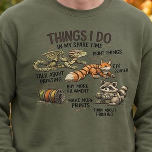 Op de afbeelding: Een olijfgroen sweatshirt met een opdruk "THINGS I DO IN MY SPARE TIME" en illustraties van een draak, een vos en een wasbeer. Het ontwerp bevat de teksten "PRINT THINGS", "FIX PRINTER", "TALK ABOUT PRINTING", "BUY MORE FILAMENT", "MAKE MORE PRINTS" en "THINK ABOUT PRINTING" bij een spoel.