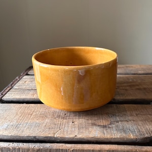May include: A round, ceramic bowl with a glossy, golden-yellow glaze. The bowl has a simple, cylindrical shape and sits on a wooden surface. The interior of the bowl is visible, and the lighting creates highlights and shadows on the surface.