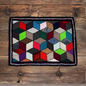 May include: A colorful, geometric rug featuring a 3D cube pattern in various colors including red, green, blue, and gray. The rug has a black border with a thin, multi-colored trim. The rug is rectangular and appears to be handmade.