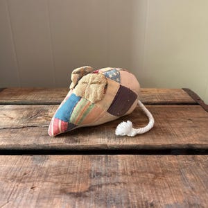 May include: A heart-shaped, patchwork fabric doorstop with a white rope tail. The doorstop features a variety of fabric patterns, including blue, red, and beige. The doorstop is sitting on a wooden surface.