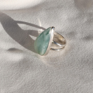 May include: A silver ring with a large, teardrop-shaped, pale green stone. The ring has a double band design and is set against a white, textured background. The stone has a smooth, polished surface.