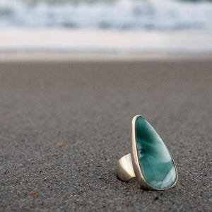 May include: A silver ring with a large, teardrop-shaped, turquoise stone. The ring is set on a sandy beach with the ocean in the background. The stone has a marbled appearance with white and blue hues.