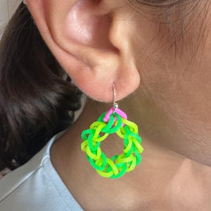 May include: A bright green and yellow woven earring with a pink detail. The earring is circular and attached to a silver hook. The earring is worn on a person's ear.
