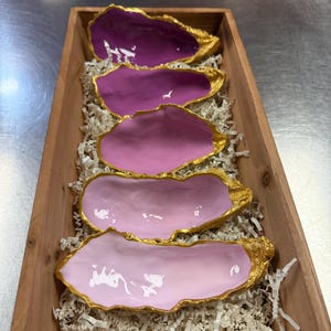 May include: A wooden tray filled with five oyster shell dishes. The shells are painted in shades of pink and purple, with gold leaf edging. The tray is filled with shredded paper for presentation.