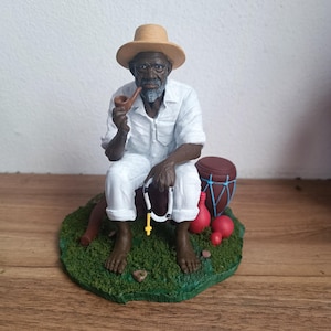 Peut inclure: Figurine d'un vieil homme noir portant un chapeau de paille et des vêtements blancs, tenant une pipe et un chapelet. Il est assis sur une base verte, avec un tambour et des objets rouges. La figurine est très détaillée.