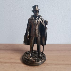 Könnte beinhalten: Bronze-farbene Figur eines Skeletts in Anzug, Zylinder und Umhang, das einen Schädelschädel-Knauf-Stock hält. Die Figur steht auf einem runden Sockel mit Knochen-Details. Die Statue ist eine detaillierte Darstellung.