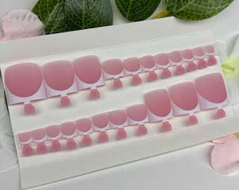 May include: Two sets of pink and white press-on toenails are displayed on a white card. The nails have a rounded shape with a pink gradient and white tips. The card is surrounded by green leaves and pink flower petals.