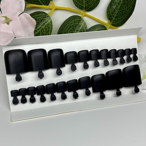 Puede incluir: Un conjunto de uñas postizas para los pies en color negro mate, dispuestas en dos filas sobre una tarjeta de presentación blanca. Las uñas varían en tamaño y cada una tiene una pequeña pestaña en la base para su aplicación. Están numeradas.