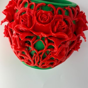 May include: A cylindrical green container featuring an intricate, raised red overlay of sculpted roses and swirling vine patterns. The vibrant red floral design wraps around the exterior of the green vessel, creating a textured, three-dimensional surface.