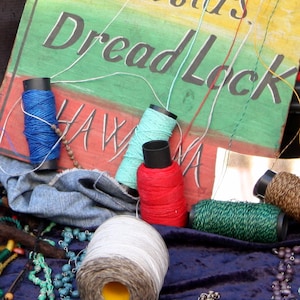 May include: A close-up of a variety of colorful spools of thread, including blue, green, red, and brown. The spools are arranged on a blue velvet surface with a sign that reads "Rastas Dread Lock" in black lettering. The sign is painted on a yellow, green, and red background.
