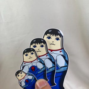 May include: A set of four nesting dolls with a blue, white, and red color scheme. The dolls have a cartoon-like design with black hair and blue eyes. The dolls are held by a hand against a white background.