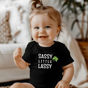 May include: A smiling baby wearing a black t-shirt with the words "SASSY LITTLE LASSY" in white, and a green shamrock. The baby has blonde hair and is sitting in front of a blurred background.