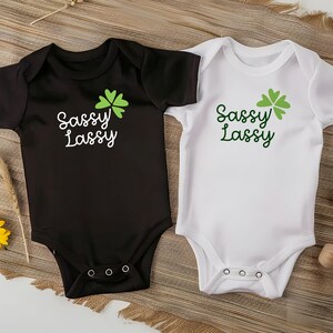 May include: Two baby onesies, one black and one white, with the text "Sassy Lassy" and a green shamrock graphic. The onesies are short-sleeved and have three snaps at the bottom. The background includes yellow flowers and a woven mat.