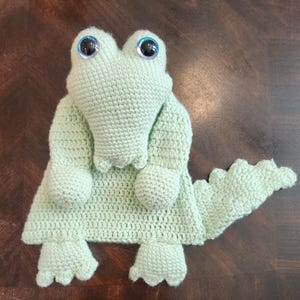 May include: A light green crocheted alligator toy with large blue eyes. The handmade reptile has a detailed snout, arms, legs, and a textured tail. The toy is made of soft yarn and is designed to be a plush toy.