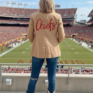 May include: Tan blazer with the word "Chiefs" in red script on the back. The person is wearing blue jeans and red cowboy boots. The background shows a football stadium filled with people.