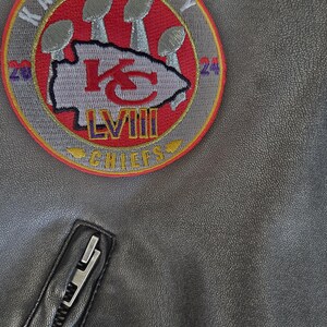 以下が含まれることがあります： グレーのジャケットのクローズアップ。円形のパッチには「Kansas City Chiefs」の文字とチームロゴが描かれています。パッチにはローマ数字の「LVIII」も表示されています。黒いジッパーが見えます。