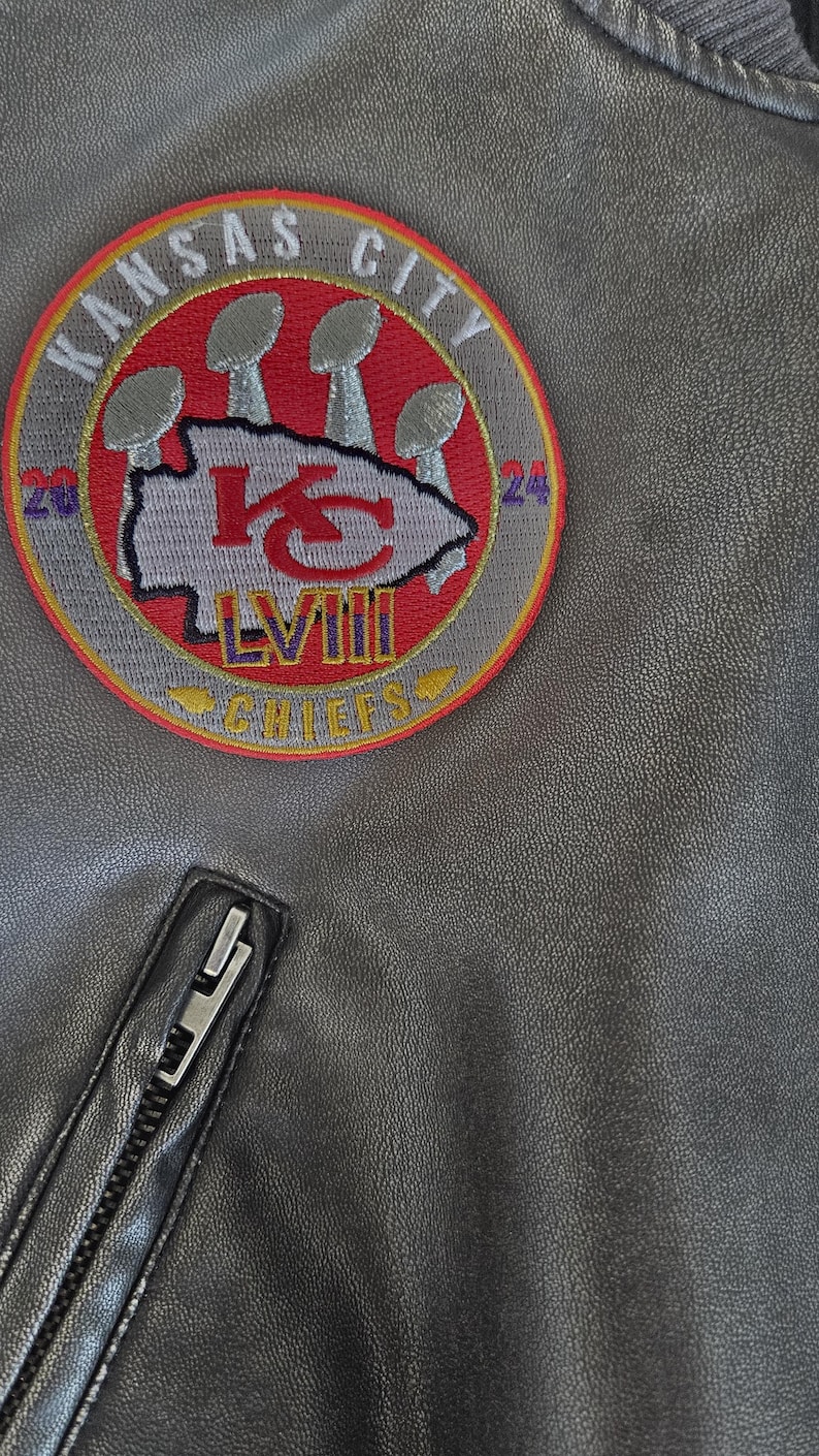 以下が含まれることがあります： グレーのジャケットのクローズアップ。円形のパッチには「Kansas City Chiefs」の文字とチームロゴが描かれています。パッチにはローマ数字の「LVIII」も表示されています。黒いジッパーが見えます。