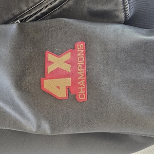 以下が含まれることがあります： 濃いグレーの革製ジャケットの袖のクローズアップ。 「4X CHAMPIONS」の文字が入った赤と金のパッチが袖に縫い付けられています。 ジャケットはリブ編みの濃いグレーのカフとジッパーが付いています。
