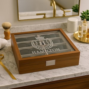 Puede incluir: Un organizador de gafas de sol de madera con tapa de cristal transparente. La caja está grabada con un diseño de escudo y el nombre "HAMILTON". El interior está dividido en compartimentos para guardar varios pares de gafas de sol.