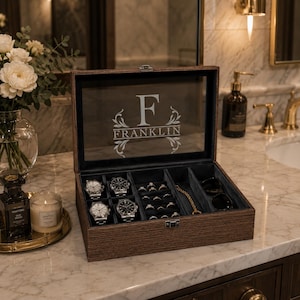 May include: A dark brown wooden watch and jewelry storage box with a glass lid featuring the engraved letter F and the name FRANKLIN. The interior is lined with dark fabric and contains four wristwatches, several rings, a gold chain, and sunglasses.
