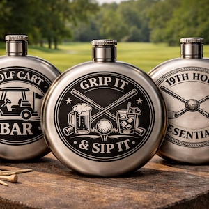 Puede incluir: Tres petacas de acero inoxidable con diseños temáticos de golf. Una petaca dice "Golf Cart Bar", otra dice "Grip It & Sip It", y la tercera dice "19th Hole Essentials". Pelotas y tees de golf en primer plano.