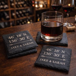 May include: A set of four square slate coasters with personalized text. Each coaster has the coordinates "46° 18' N 136° 30' E" and the names "JAKE & SARAH" etched in white. A glass of red wine sits on one coaster.