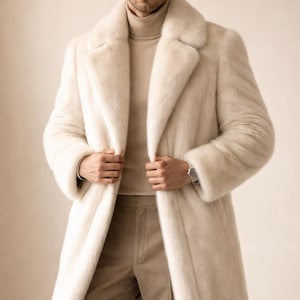 May include: A long, cream-colored fur coat with a wide collar. The coat is worn over a beige turtleneck and matching trousers. The model is wearing a silver wristwatch. The coat appears to be made of a soft, luxurious material.