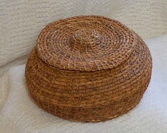 May include: A round, lidded basket woven from natural-colored fibers. The basket's body and lid are constructed with a tight, spiral weave, creating a textured surface. The lid fits snugly on top, and the basket appears to be handmade.