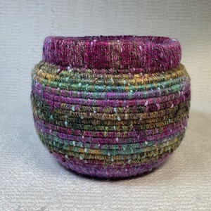 Puede incluir: Una cesta redonda tejida a mano con una abertura ancha. La cesta presenta una paleta de colores púrpura, verde y dorado, con motas blancas. El borde es de un tono púrpura más oscuro. Esta cesta decorativa es adecuada para el almacenamiento.
