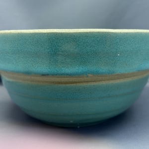 May include: A teal ceramic bowl with a textured surface. The bowl has a wide rim and a slightly flared shape. The exterior is a solid teal color, while the rim has a lighter, off-white hue. The bowl is suitable for serving food.