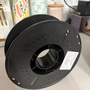 Puede incluir: Una bobina de filamento negra para impresora 3D, con un agujero central y numerosas perforaciones pequeñas. La bobina, hecha de un material oscuro, está diseñada para contener filamento para la impresión 3D. Una etiqueta es parcialmente visible.