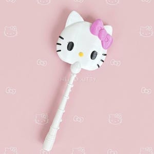 May include: A white plastic wand featuring a Hello Kitty character head at the top. The character has black oval eyes, a yellow nose, and a pink bow on the right side. The handle is white with a segmented, ribbed design extending downwards.