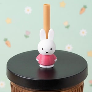 Op de afbeelding: Een rietjes-topper met een wit konijnfiguurtje met lange oren en een zwarte X-vormige mond, gekleed in een effen roze jurk. De topper is geplaatst op een beige rietje dat door een zwart deksel op een geribbelde beker steekt.
