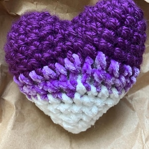 May include: A handmade, plush heart in shades of purple and white. The top half is a deep purple, transitioning to lighter purple and then white at the bottom. The heart is made of soft, textured yarn, perfect for a cozy gift or decoration.