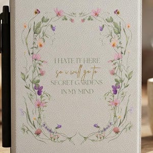 May include: A cream-colored e-reader case featuring a delicate floral wreath design surrounding the text: I HATE IT HERE so i will go to SECRET GARDENS IN MY MIND. The case includes a black stylus holder on the left side and a textured finish.