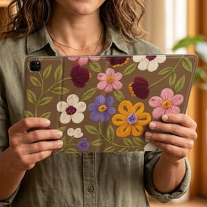 May include: A tablet case featuring a brown background with a vibrant floral pattern. The design includes various stylized flowers in shades of pink, purple, orange, and white, accompanied by green leaves and stems on a brown base.