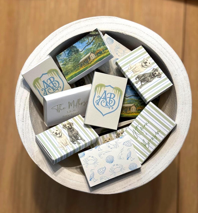 May include: A collection of decorative matchboxes in a wooden bowl. The matchboxes feature various designs, including a house, dogs, and a monogram. Some boxes have striped patterns and text.