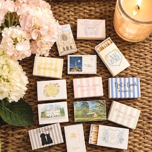 May include: A collection of decorative matchboxes with various designs and text, including floral patterns, architectural illustrations, and personalized names and dates. The matchboxes are arranged on a woven surface, with a lit candle and flowers in the background.
