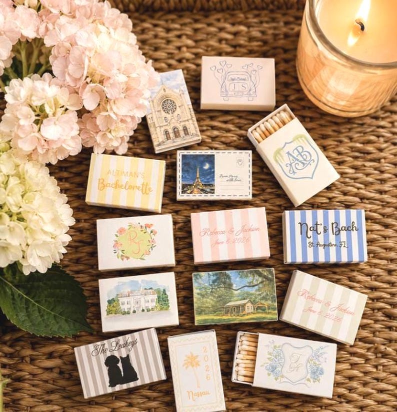 May include: A collection of decorative matchboxes with various designs and text, including floral patterns, architectural illustrations, and personalized names and dates. The matchboxes are arranged on a woven surface, with a lit candle and flowers in the background.