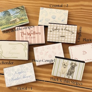May include: A collection of decorative matchboxes with various designs. The boxes feature themes like coastal scenes, floral patterns, and text such as "The Walkers" and "Kaylee's Bridal Shower." Some boxes have illustrations of dogs, seashells, and a coffee cup.