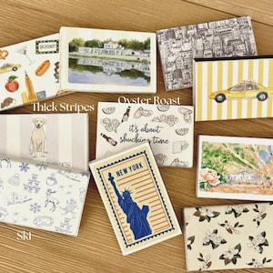 May include: A collection of decorative matchboxes with various designs. One box features a yellow taxi, another has a dog, and others display cityscapes, food, and the Statue of Liberty. The boxes are rectangular and contain wooden matches.