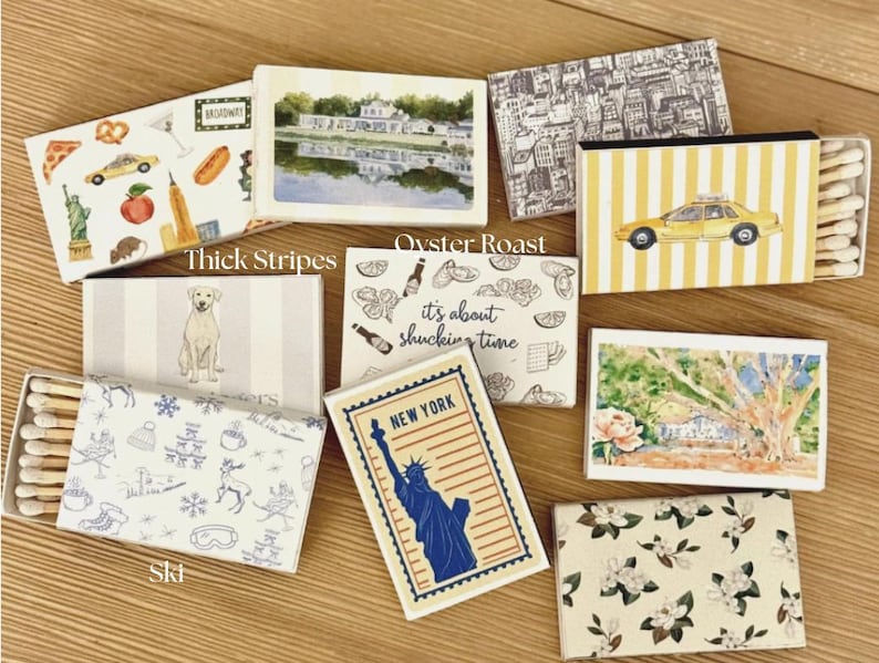 May include: A collection of decorative matchboxes with various designs. One box features a yellow taxi, another has a dog, and others display cityscapes, food, and the Statue of Liberty. The boxes are rectangular and contain wooden matches.