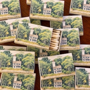 May include: A collection of matchboxes featuring a watercolor painting of a lighthouse and a house nestled among lush green trees. One box is open, revealing the wooden matchsticks inside. The boxes are arranged on a dark wooden surface.