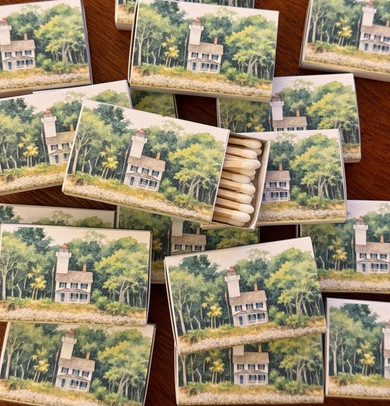 May include: A collection of matchboxes featuring a watercolor painting of a lighthouse and a house nestled among lush green trees. One box is open, revealing the wooden matchsticks inside. The boxes are arranged on a dark wooden surface.